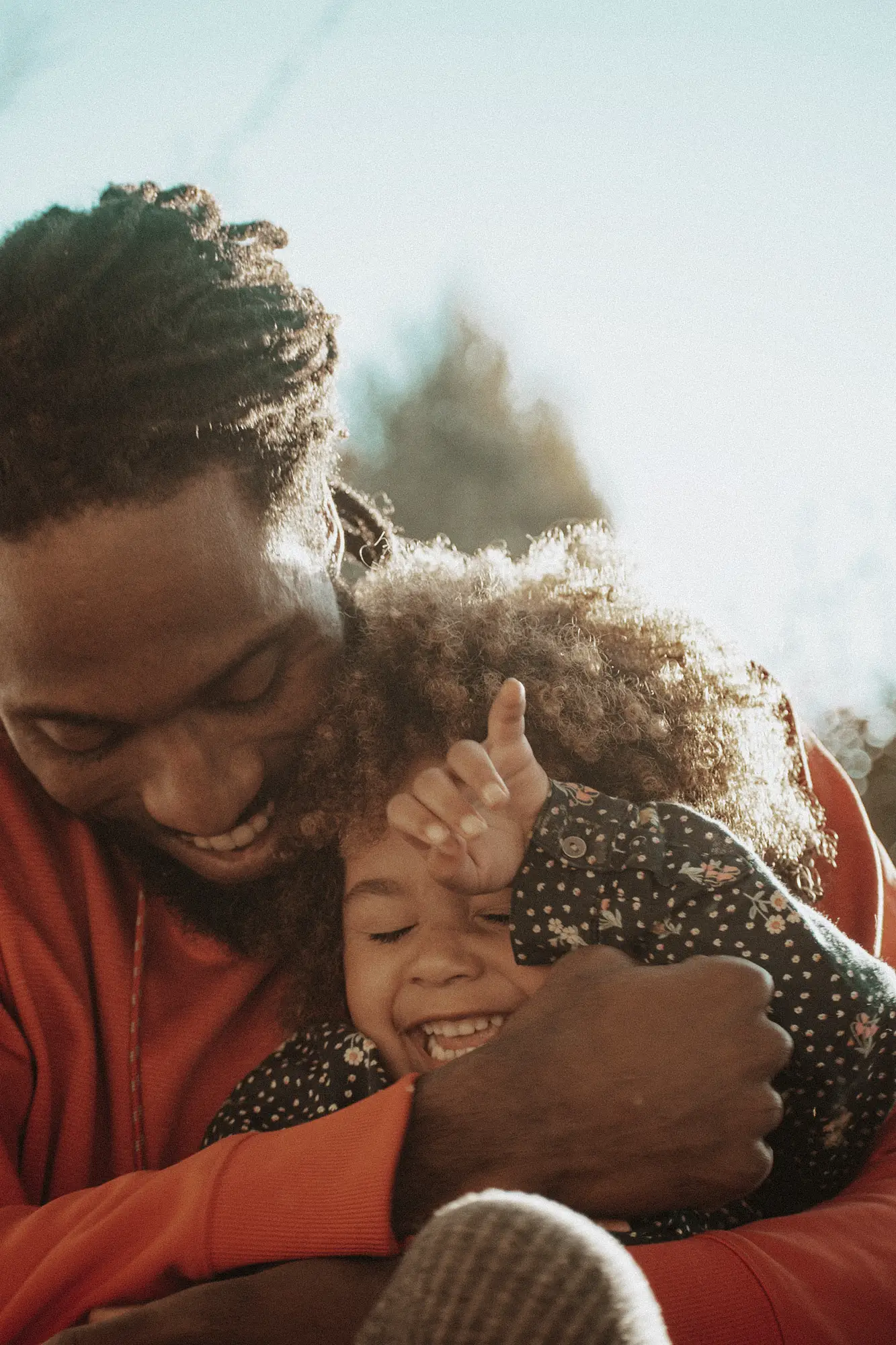 Vater umarmt lachendes Kind mit lockigem Haar im Freien bei Sonnenlicht.