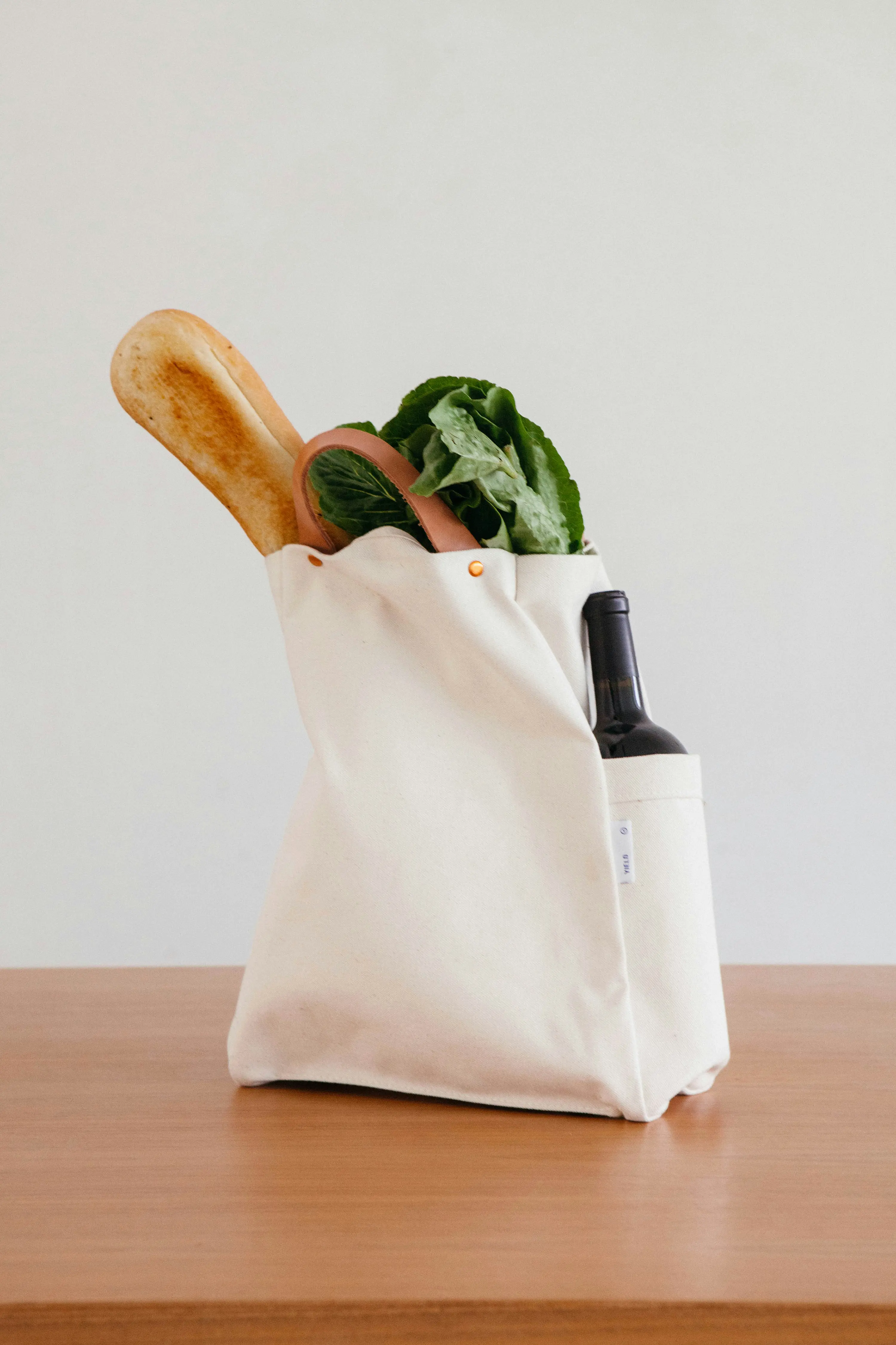 Leinentasche mit einem Baguette, grünem Salat und einer Weinflasche auf einem Holztisch.