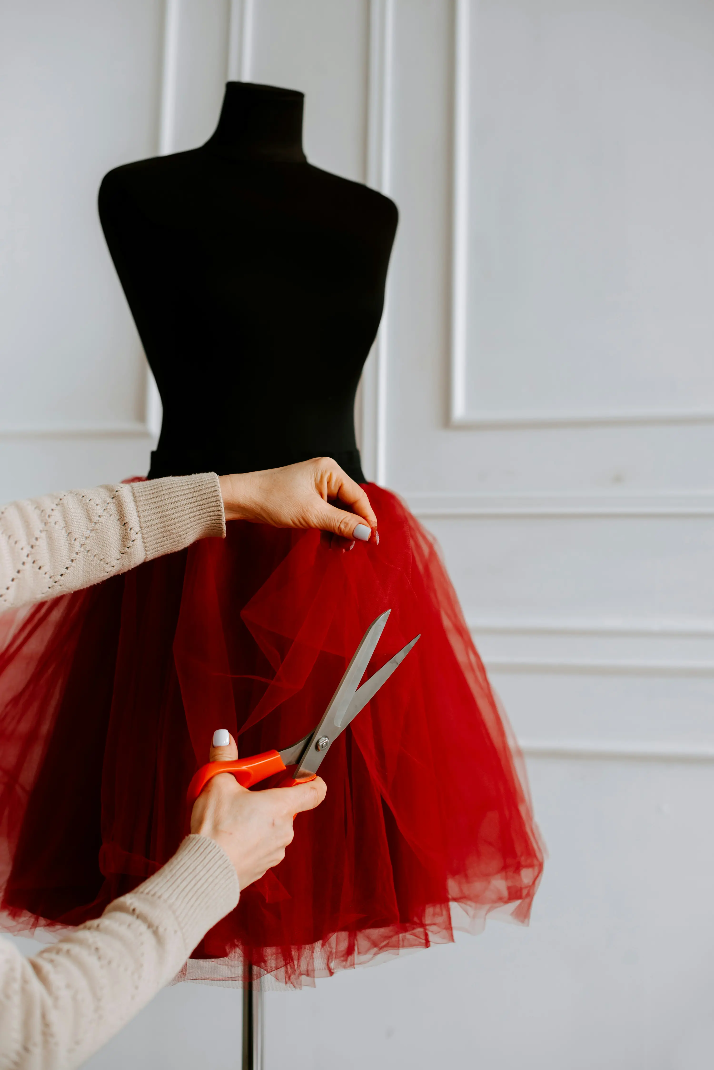 Person cuts red tulle fabric on a black dress form with orange scissors in a bright room.